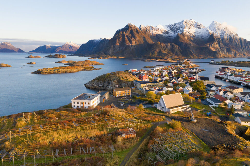 Henningsvær i Lofoten med fargerike hus og snødekte fjell ved havet, under en klar himmel.
